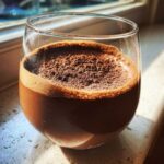 A close-up of rich, airy chocolate mousse topped with cocoa powder, served in a clear glass near a window.