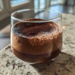 A close-up of airy, rich chocolate mousse dusted with cocoa powder in a clear glass.