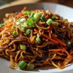 A close-up shot of glossy, savory fried noodles mixed with julienned carrots and topped with fresh green onions.