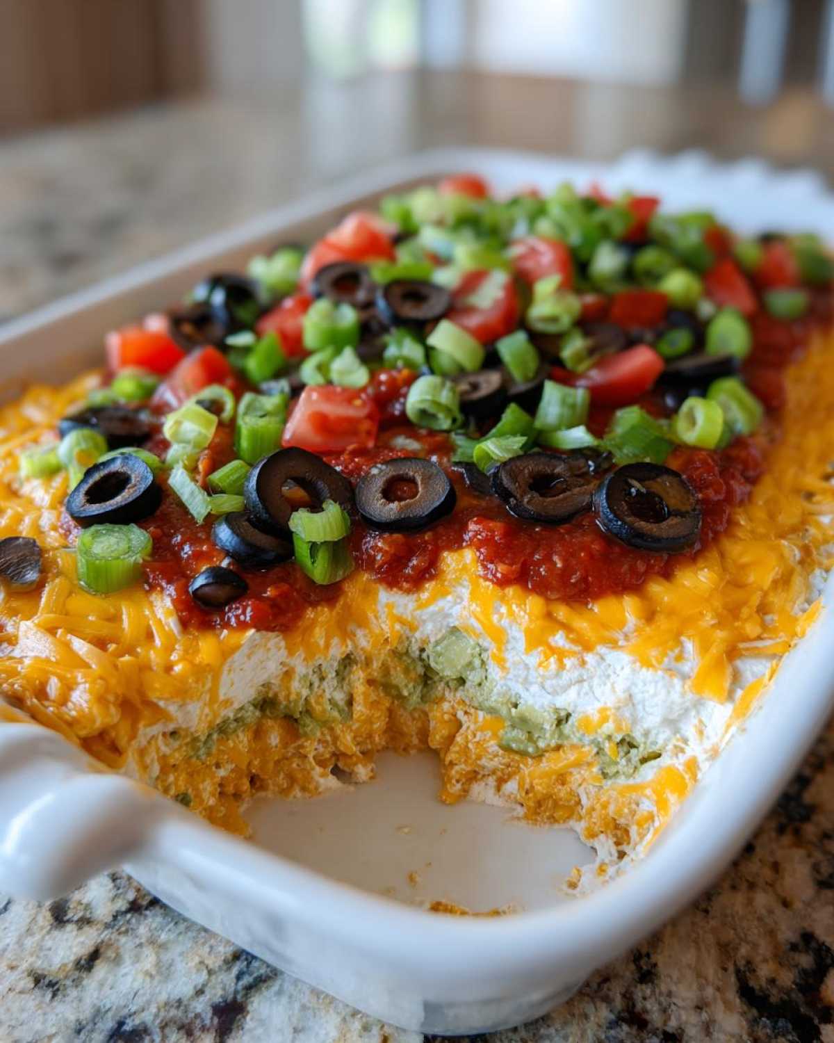 A close-up view of a vibrant 7 layer dip in a white rectangular dish, showing layers of cheese, guacamole, sour cream, salsa, olives, and green onions.