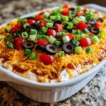 A vibrant close-up of a fully assembled 7 layer dip topped with shredded cheese, black olives, tomatoes, and green onions.