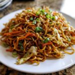A close-up of a generous serving of glossy, brown fried noodles mixed with shredded carrots and cabbage, topped with fresh green onions.