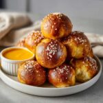 A stack of warm, golden brown pretzel bites topped with coarse salt, served next to a small bowl of bright yellow cheese sauce.