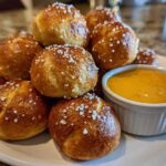A pile of freshly baked, golden brown pretzel bites topped with coarse salt, served with a side of creamy cheese sauce.