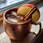 A frosty copper mug filled with an Apple Cider Moscow Mule, garnished with apple slices and cinnamon sticks.