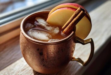 A frosty copper mug filled with an Apple Cider Moscow Mule, garnished with apple slices and cinnamon sticks.