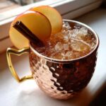 Close-up of a chilled apple cider moscow mule in a hammered copper mug, garnished with apple slices and a cinnamon stick.