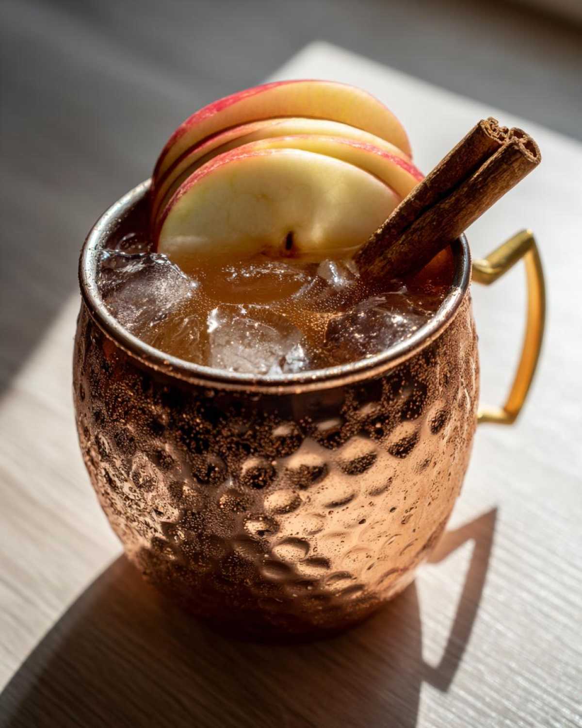 A chilled apple cider moscow mule garnished with thin apple slices and a cinnamon stick in a hammered copper mug.