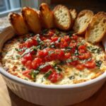 A white baking dish filled with hot, cheesy bruschetta dip topped with fresh tomatoes and basil, served with toasted bread slices.