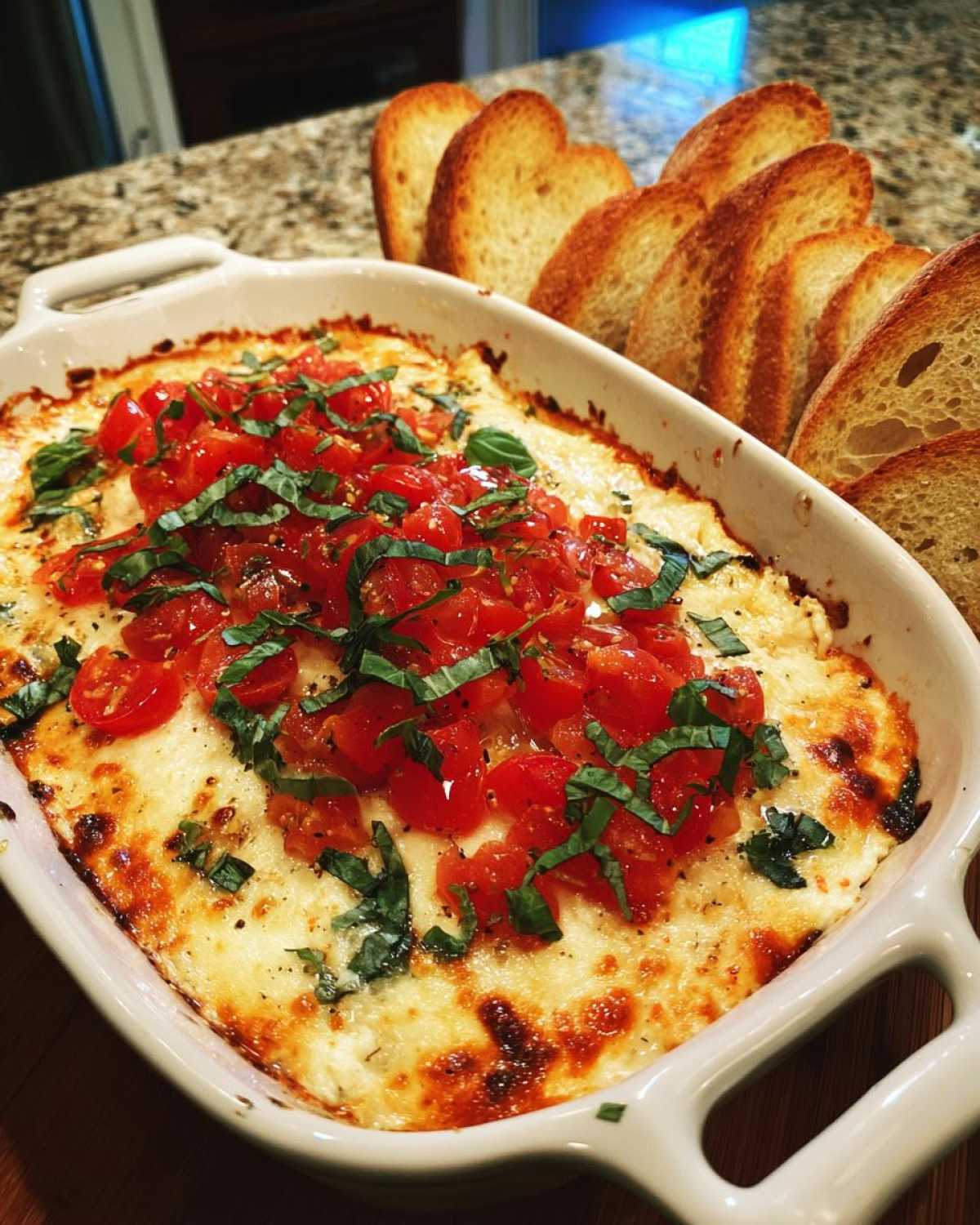 Baked bruschetta dip in a white casserole dish, topped with diced tomatoes and fresh basil, served with toasted bread.