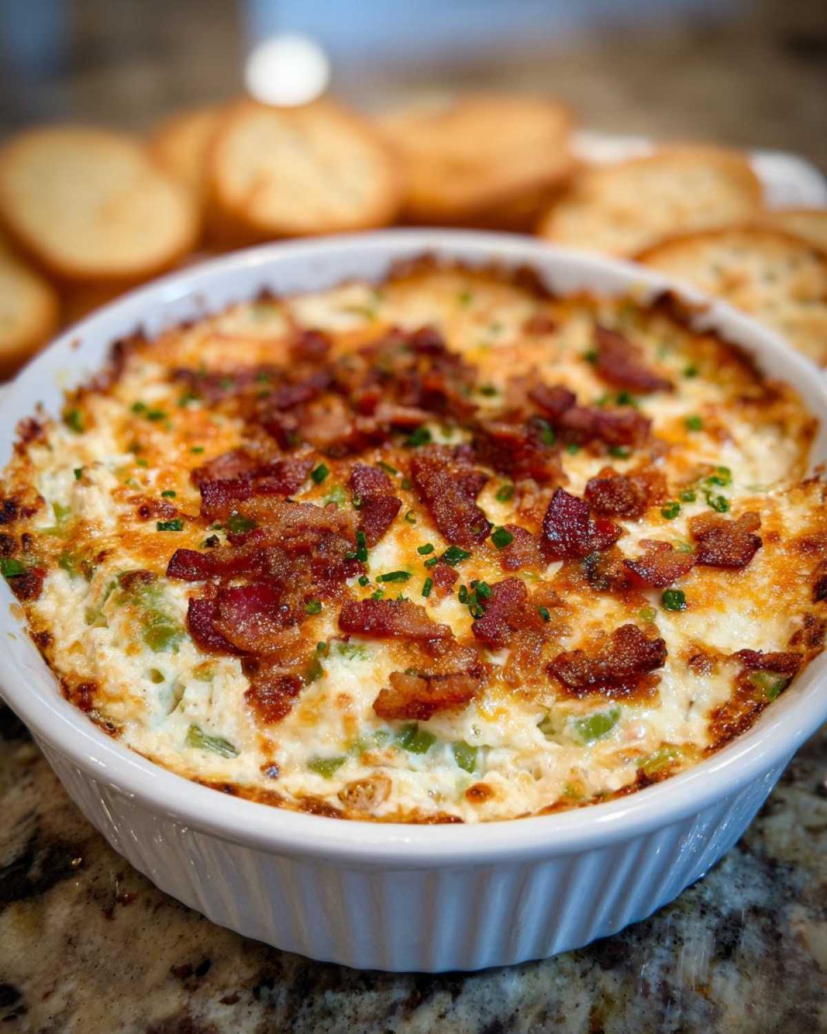 A close-up of hot baked jalapeño popper dip in a white dish, topped with crispy bacon bits and chives.