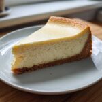 A perfect slice of creamy baked cheesecake recipe with a thick graham cracker crust on a white plate.