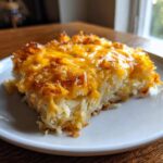 A square serving of cheesy potato casserole made with shredded potatoes and melted cheddar cheese.