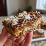 A hand holding a thick, chewy magic cookie bars square showing layers of crust, caramel, chocolate, and toasted coconut.