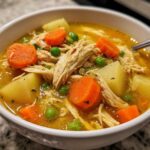 Close-up of a bowl of rich, golden chicken vegetable soup filled with shredded chicken, carrots, potatoes, and peas.