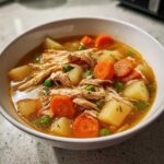 Close-up of a bowl of rich chicken vegetable soup filled with shredded chicken, carrots, potatoes, and peas.