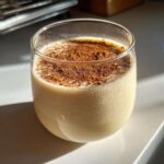 A chilled glass of creamy, pale yellow coquito topped generously with ground cinnamon.