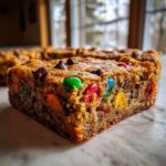 A thick, chewy slice of christmas cookie bars loaded with colorful M&Ms and chocolate chips on a marble surface.