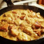 Close-up of creamy chicken and dumplings simmering in a dark cast iron skillet, featuring browned chicken pieces.