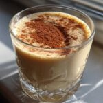A glass of creamy coquito topped generously with ground cinnamon, sitting in bright sunlight.