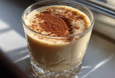 A glass of creamy coquito topped generously with ground cinnamon, sitting in bright sunlight.