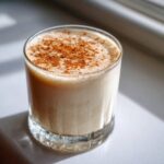 A glass of creamy coquito recipe topped generously with ground cinnamon, sitting in bright sunlight.