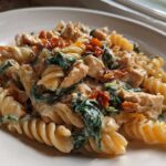 Close-up of creamy marry me chicken pasta featuring fusilli, chicken pieces, spinach, and sun-dried tomatoes on a white plate.