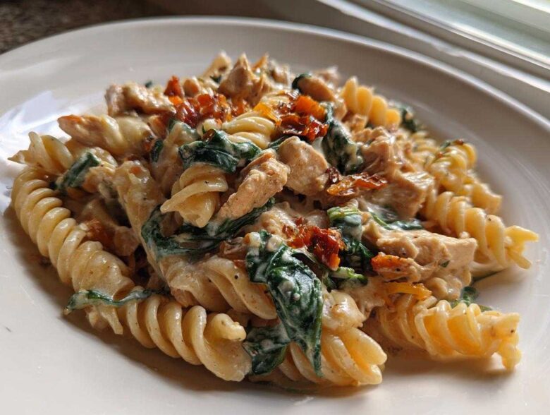 Close-up of creamy marry me chicken pasta featuring fusilli, chicken pieces, spinach, and sun-dried tomatoes on a white plate.
