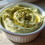 Close-up of a smooth, pale green olive dip topped with chopped pistachios and a drizzle of olive oil.