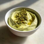 Close-up of a creamy, light green olive dip topped with chopped pistachios and olive oil.