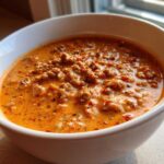 Close-up of a creamy, orange-hued pumpkin chili filled with ground meat, served in a white bowl.
