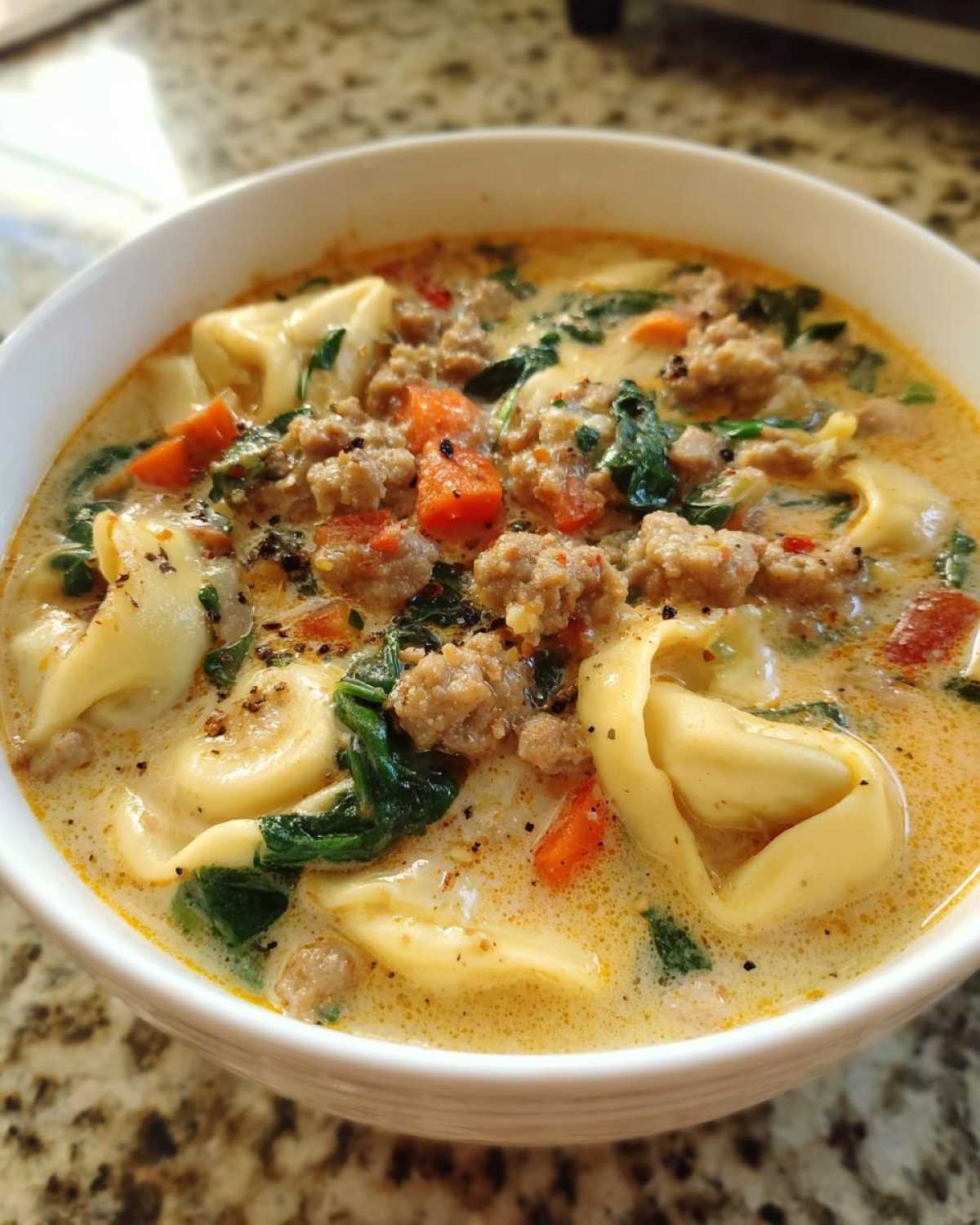 A close-up view of a hearty bowl of creamy sausage tortellini soup, filled with pasta, ground sausage, spinach, and carrots.