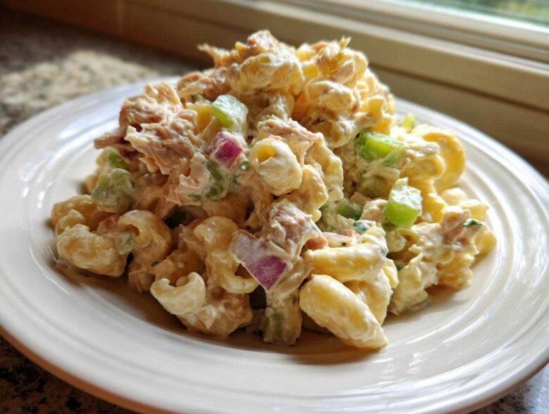 A generous serving of creamy Tuna pasta salad featuring elbow macaroni, tuna chunks, green celery, and red onion on a white plate.