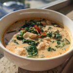 A close-up of a bowl of creamy Tuscan chicken soup featuring chunks of chicken, wilted spinach, and sun-dried tomatoes.