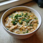 A close-up of a creamy white chicken chili in a bowl, topped generously with shredded cheese and fresh cilantro.