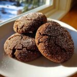 A close-up of four dark, crinkled gingersnap cookies heavily coated in sparkling sugar, resting on a white plate near a window.