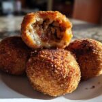 Four golden-brown, deep-fried rice balls stacked on a white plate, one cut open to show a savory meat filling inside these amazing rice balls.