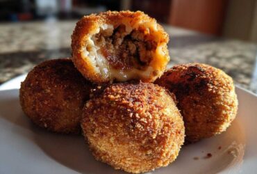Four golden-brown, deep-fried rice balls stacked on a white plate, one cut open to show a savory meat filling inside these amazing rice balls.