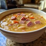 Close-up of a creamy, savory bean soup featuring white beans, diced carrots, and chunks of ham.
