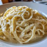 Close-up of fettuccine pasta generously coated in creamy homemade alfredo sauce and topped with grated cheese and black pepper.