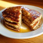 A close-up of a stack of fluffy banana pancakes, cut open to show the interior, drizzled with syrup.