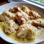 Close-up of fluffy dumplings submerged in creamy sauce with pieces of seasoned chicken in a white bowl, featuring chicken and dumplings.