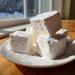 Close-up of several fluffy homemade marshmallows dusted heavily with powdered sugar on a white plate.
