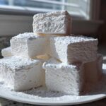 A stack of square, fluffy homemade marshmallows generously dusted with powdered sugar on a white plate.