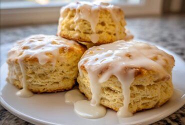 Three fluffy scones stacked slightly on a white plate, generously topped with dripping vanilla glaze.