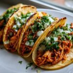 Four freshly made chicken tacos filled with shredded orange chicken, topped with diced white onion and cilantro.
