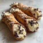 Three freshly filled cannoli with creamy ricotta filling, mini chocolate chips, and powdered sugar.