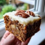 A hand holding a moist square of frosted apple brownies topped with cream cheese frosting and pecans.