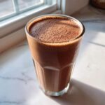 A tall glass filled with freshly made, frothy chocolate milk sitting on a white marble surface near a window.