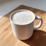 A close-up of a mug filled with creamy white hot chocolate topped with thick foam, sitting on a wooden surface.